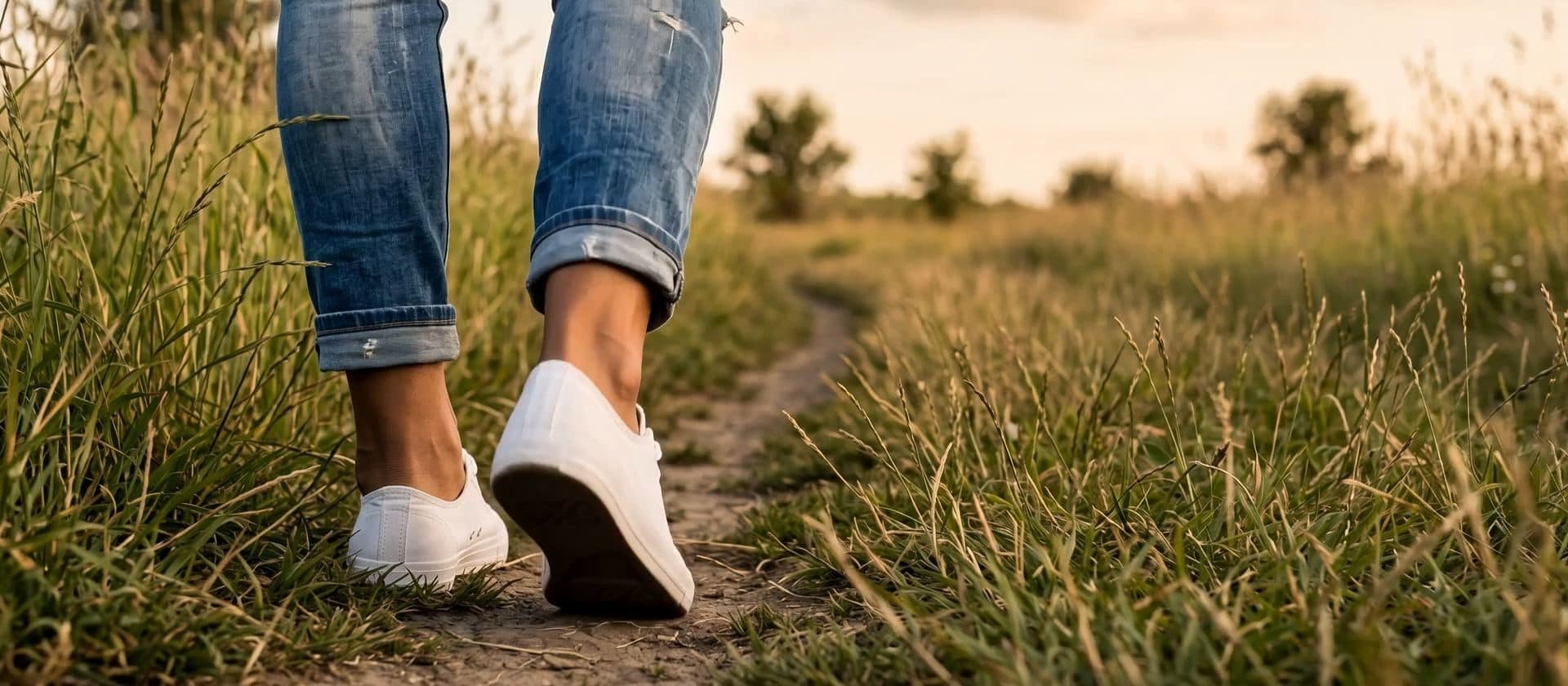 Person in jeans and white sneakers walking along a grassy path in soft golden light